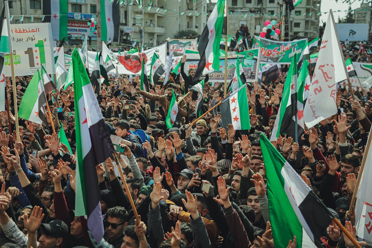 A large gathering in Syria with people holding flags and signs during a protest.